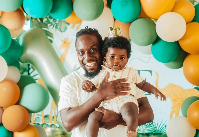A father lifts his son at his first birthday party in front of a green and orange balloon wall