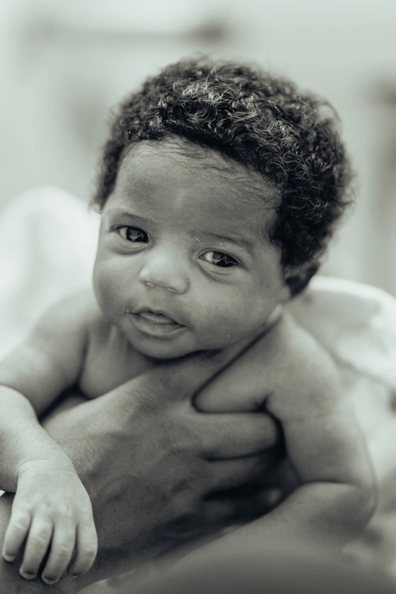 Black-and-white close portrait of a baby with curly hair, supported by an adult’s hand