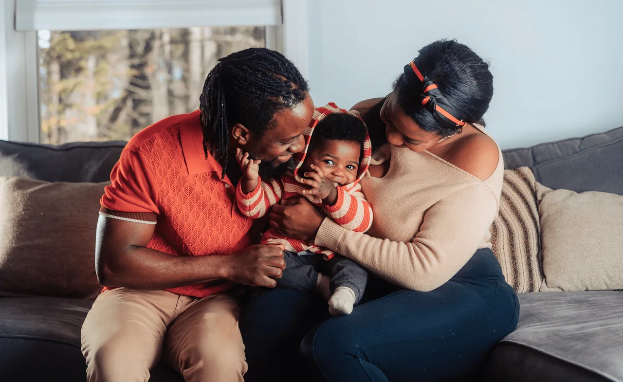 Two parents cuddle their baby between them on a gray living-room couch