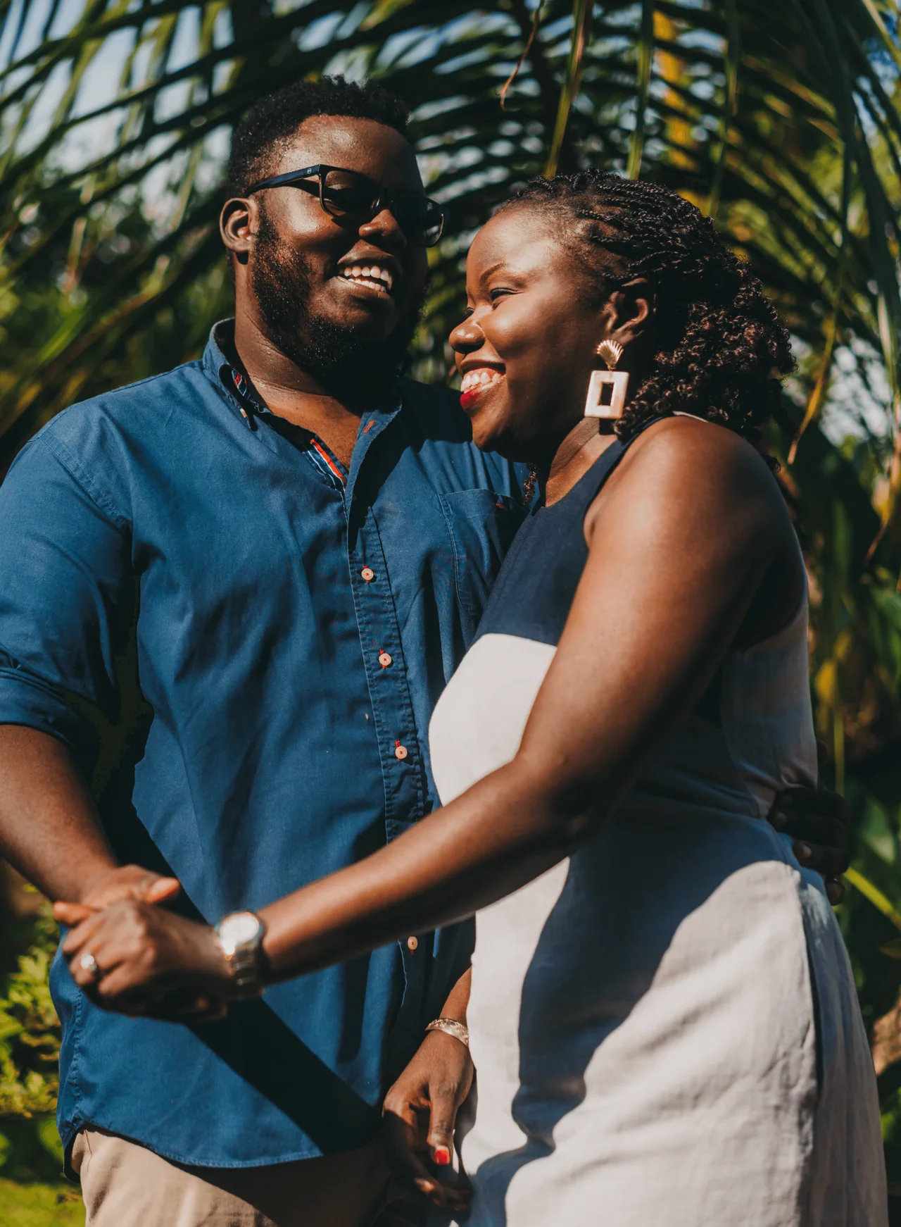 A couple smiles toward one another beneath green foliage
