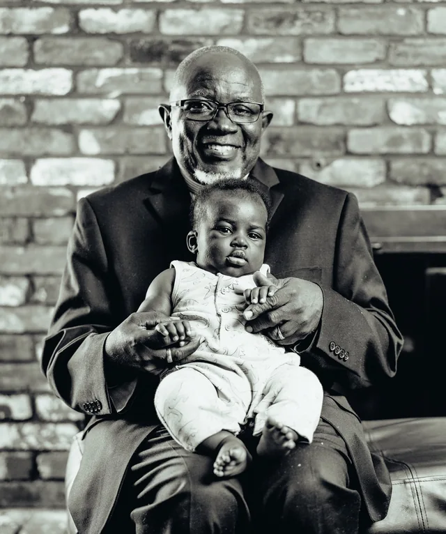 A grandfather in a suit holds his baby grandson on his lap in front of a painted brick wall