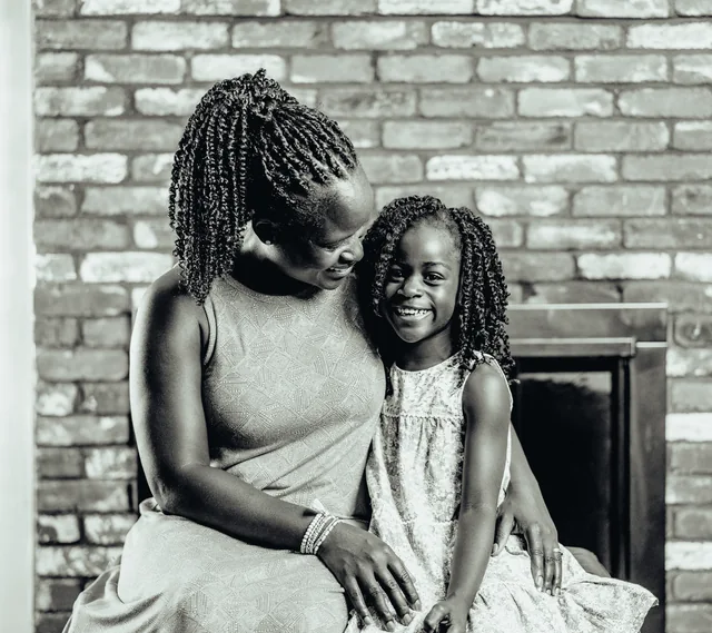 A mother and her young daughter sit cheek to cheek, both smiling, in front of a brick wall