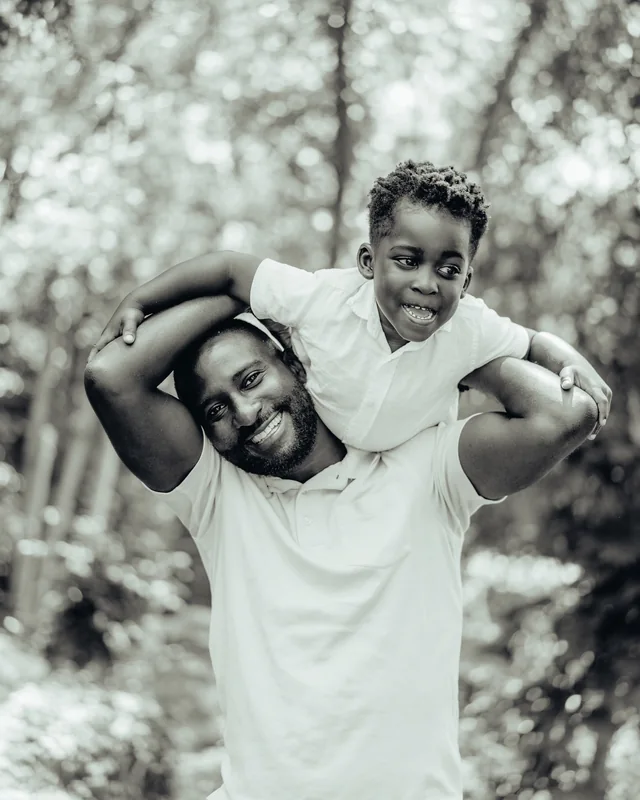 A father laughs as his young son rides on his shoulders under the trees