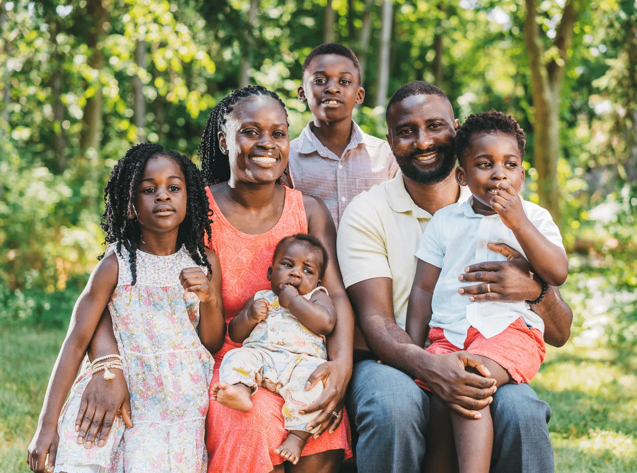 A family of six sits together on the grass in a leafy backyard, parents holding the two youngest children