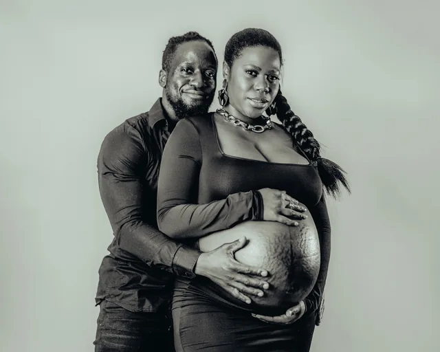 An expecting mother's partner stands behind her, hands resting on her bump
