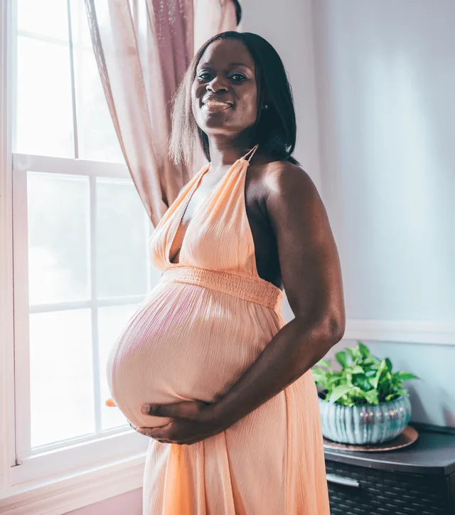 An expecting mother in a peach gown smiles by a bright window, hands on her bump