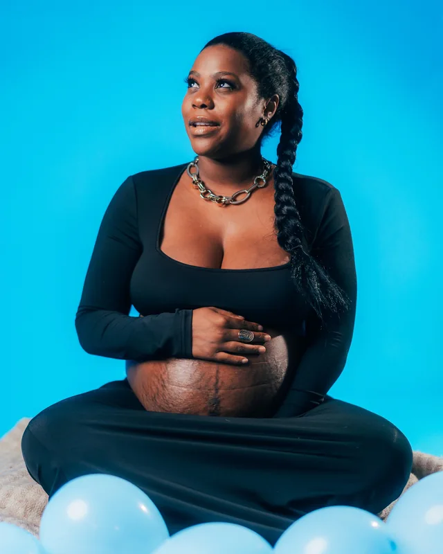 An expecting mother sits on the floor against a blue backdrop, looking up and smiling