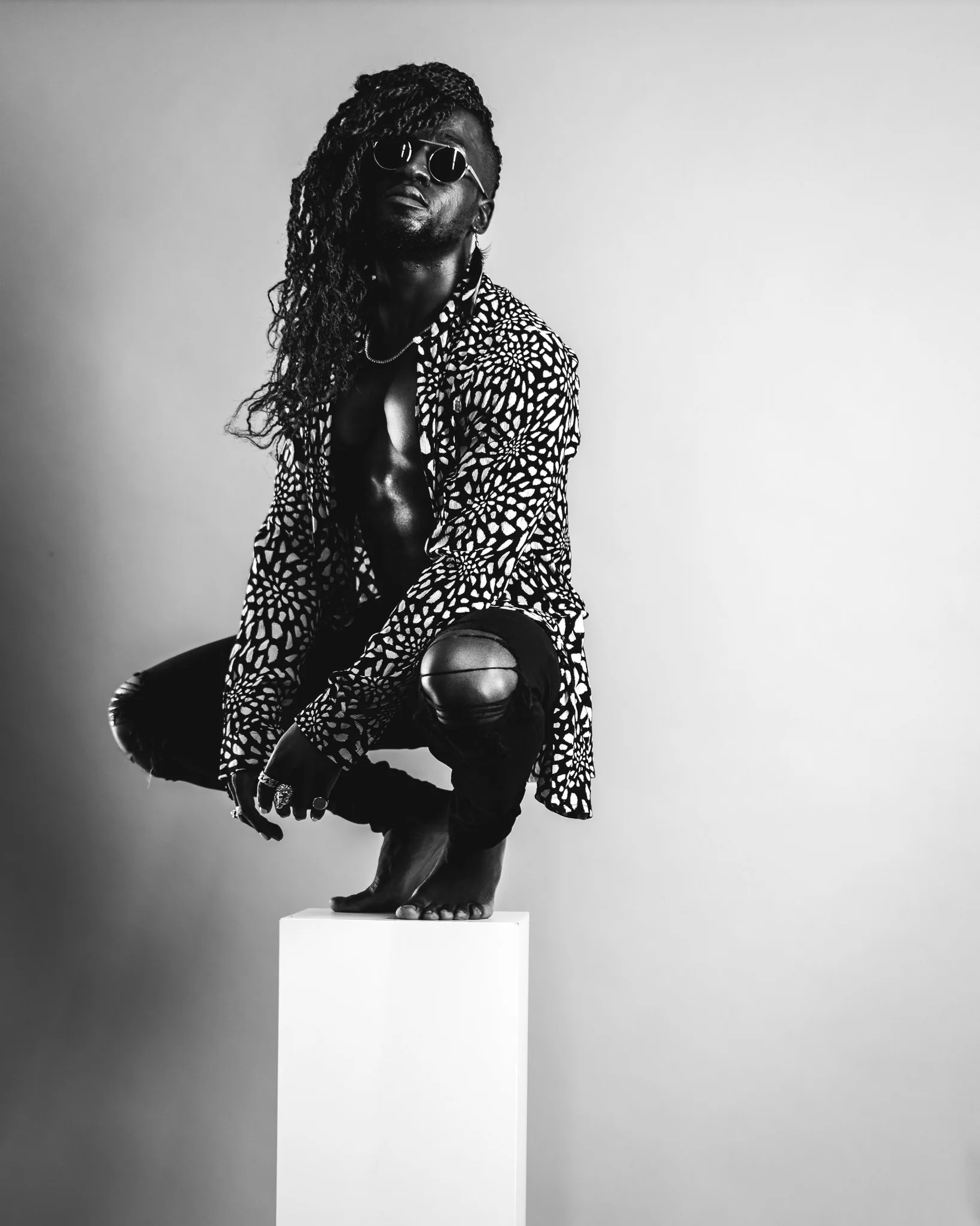 Black-and-white editorial portrait of a man in sunglasses and a patterned shirt posed on a white plinth