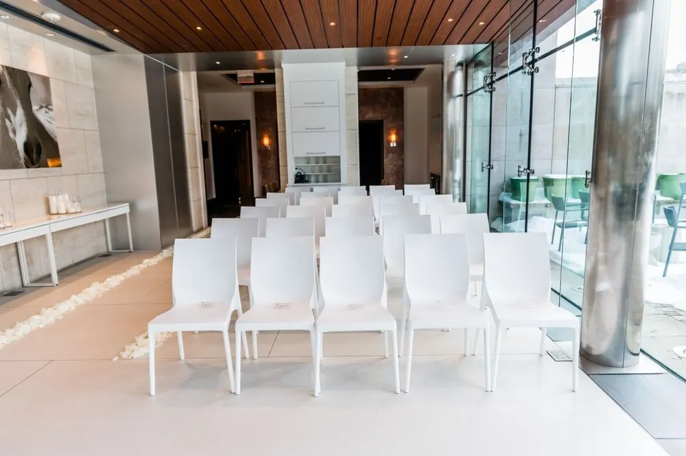 Rows of white chairs line a petal aisle beside floor-to-ceiling windows