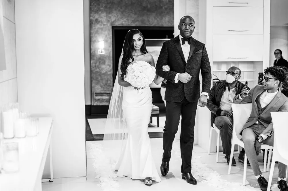 The bride walks the aisle on the arm of a man in a black brocade tuxedo, in black and white