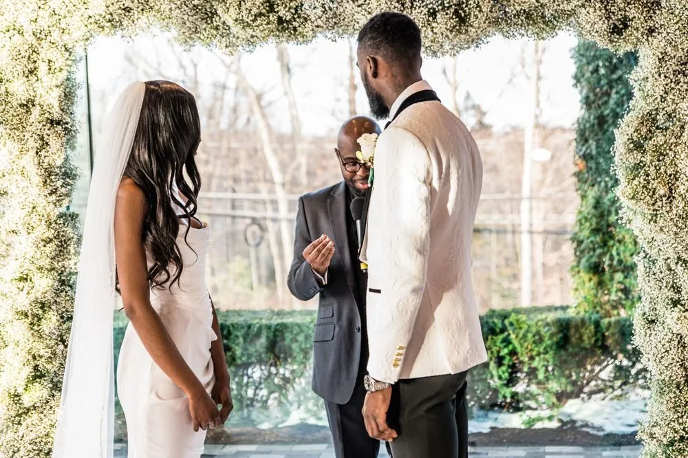 Abigail and Dontre face the officiant under the baby's breath arch with winter trees beyond the glass