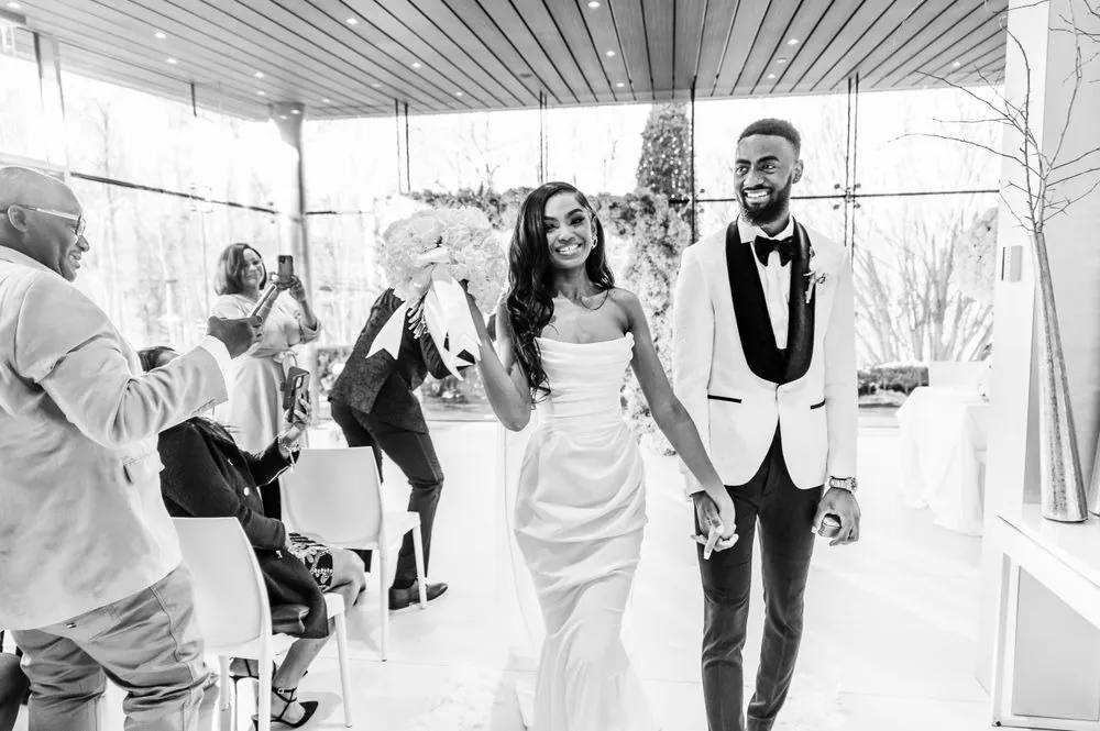Abigail and Dontre walk back up the aisle smiling as guests applaud, in black and white