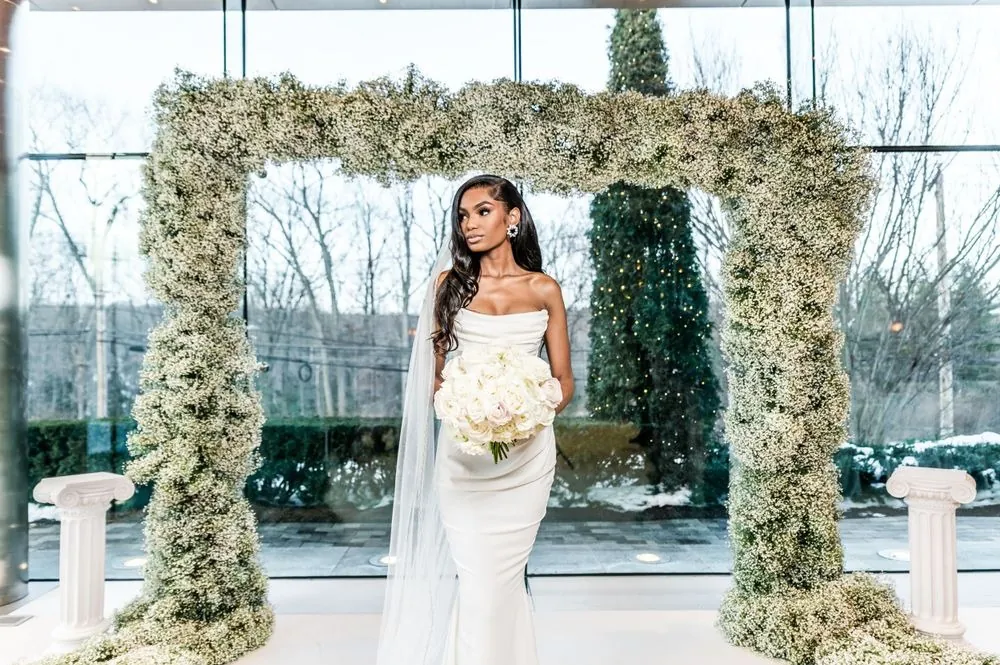 The bride stands alone beneath the arch holding a white rose bouquet