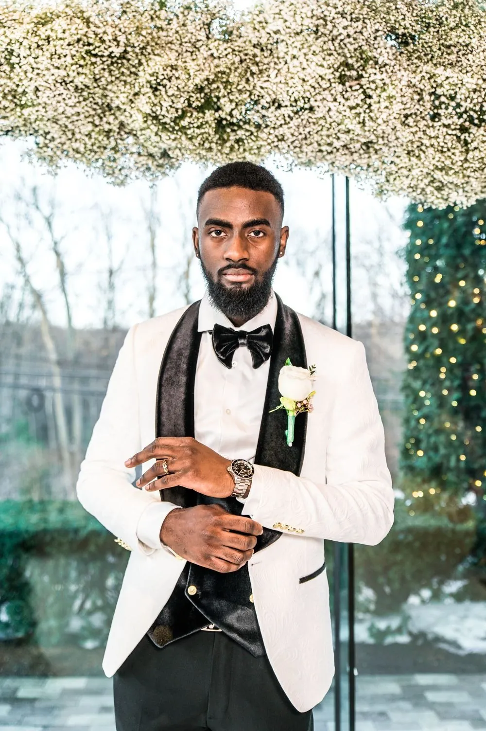 The groom in an ivory dinner jacket with black lapels stands under the floral arch