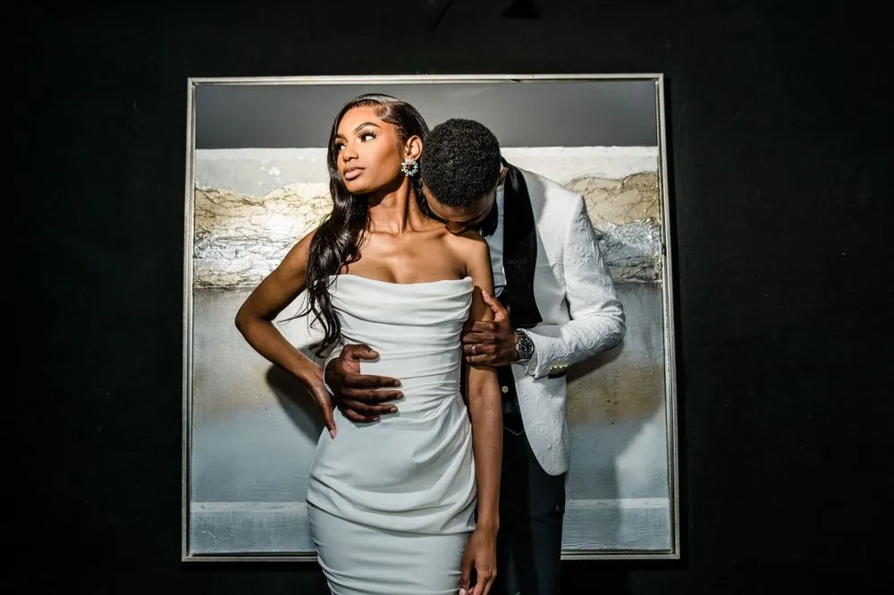 Dontre kisses Abigail's shoulder in front of a large framed painting on a dark wall