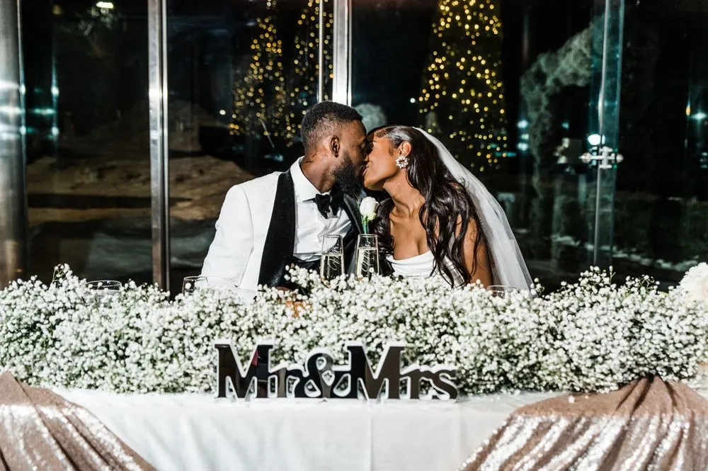 Abigail and Dontre kiss at the sweetheart table as evergreen lights glow outside
