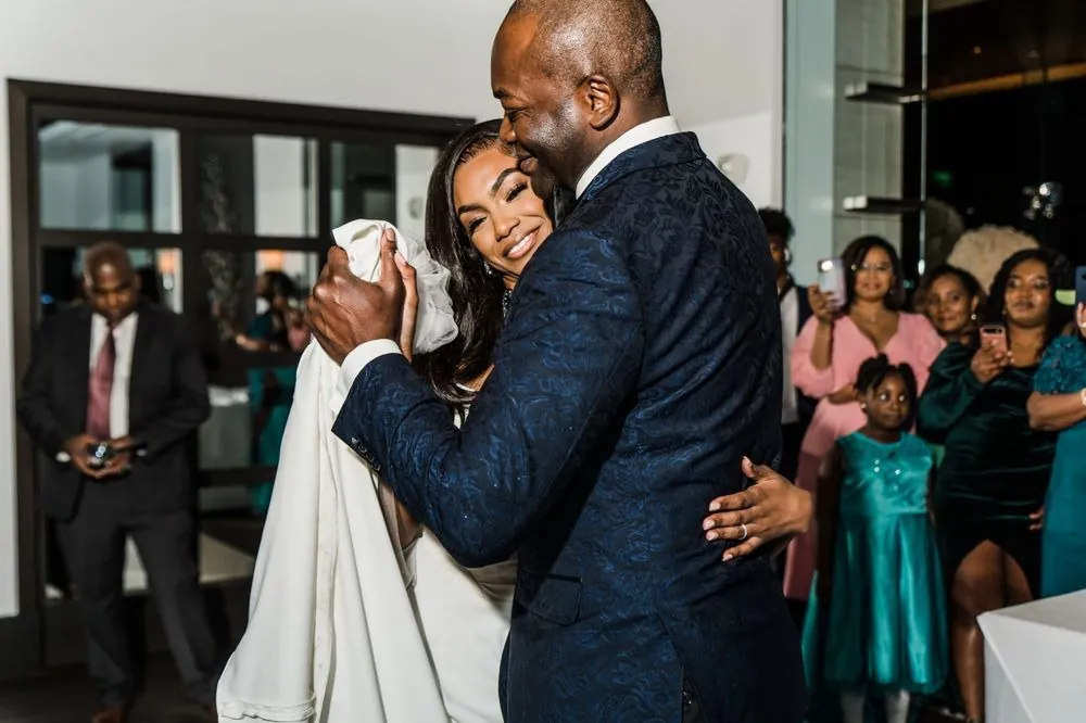 The bride dances with a man in a navy brocade jacket as family records on their phones