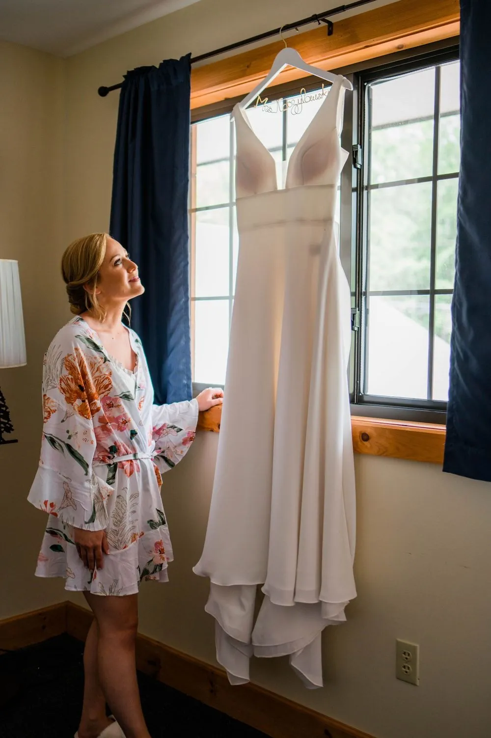 Bride in a floral robe looks up at her gown hanging in the window