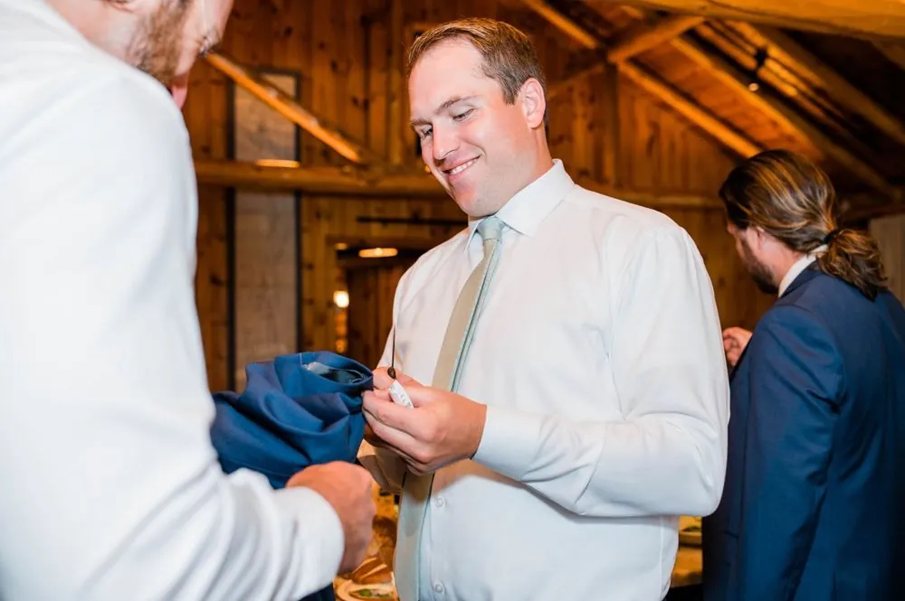 Groom in a white shirt smiles as a friend helps him dress in a wood-paneled room