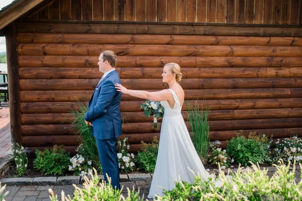 Alexandra touches Robert's shoulder from behind at a log cabin wall for their first look