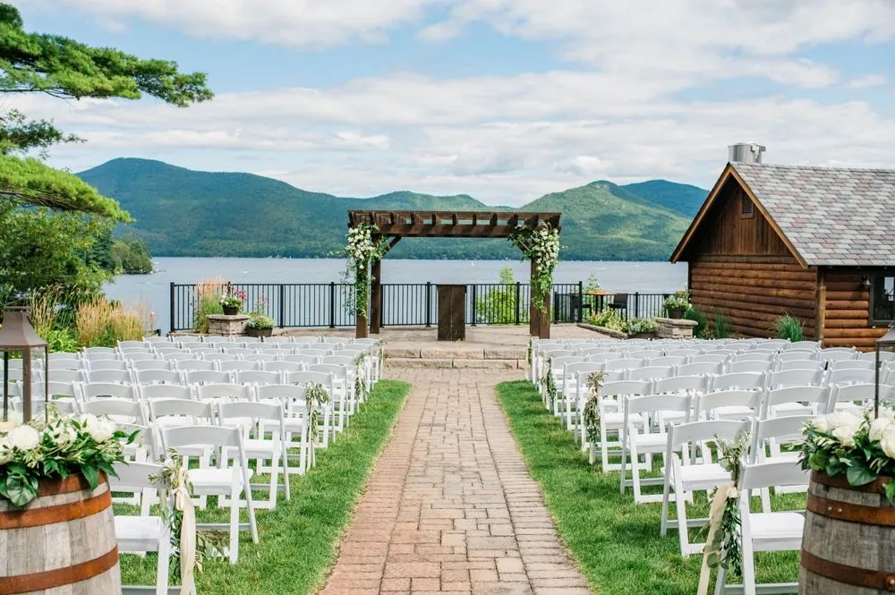 Brick aisle through white chairs leads to a greenery pergola above Lake George