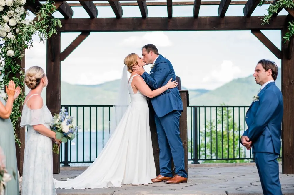 Alexandra and Robert kiss under the wooden pergola with the lake beyond the railing