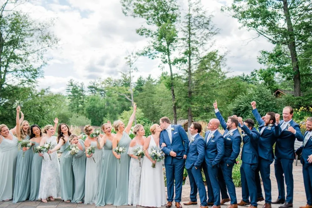 Bridesmaids in sage and groomsmen in navy cheer on the lawn among tall pines