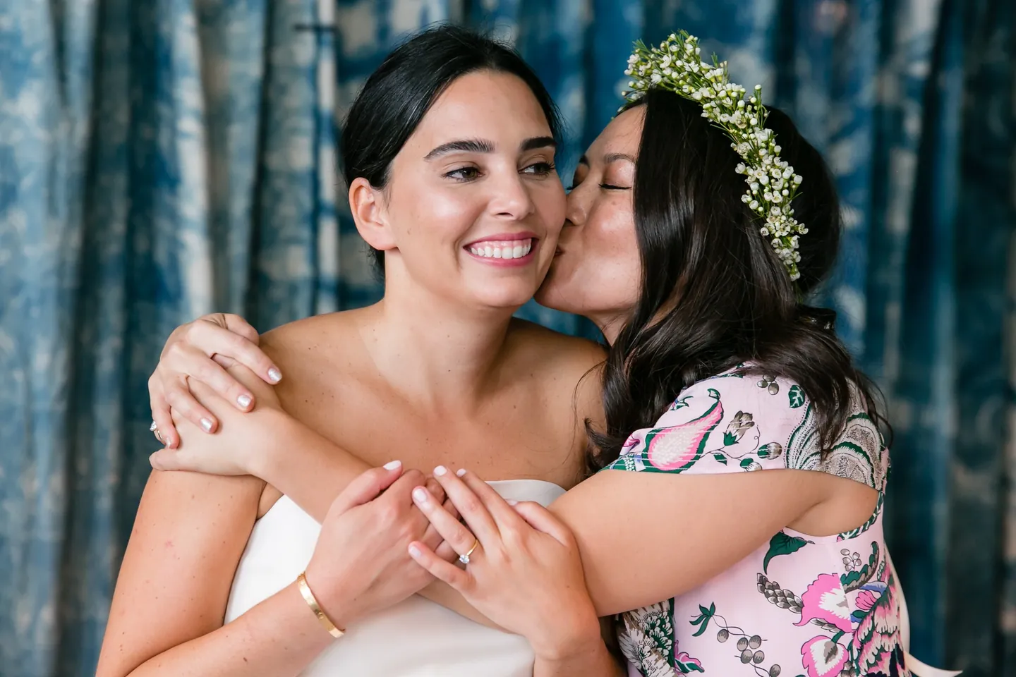 A friend in a crown of greenery kisses the bride's cheek in front of indigo curtains