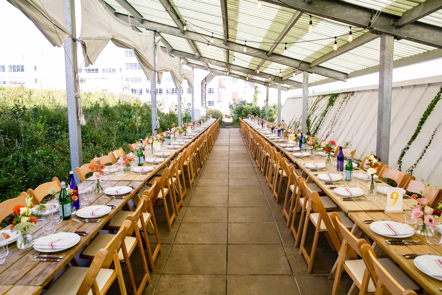 Long wooden tables set end to end beneath a pergola beside the planting beds