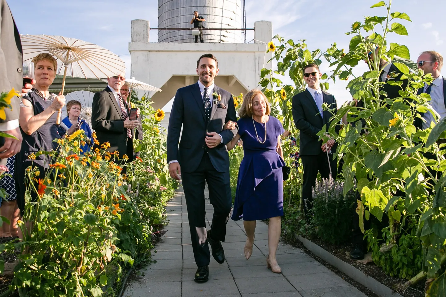The groom walks the garden path with a woman in blue as guests watch from the rows