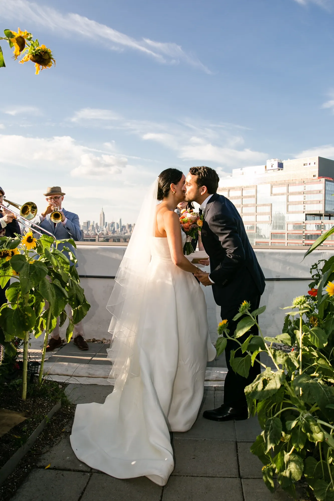 Alexis and David kiss at the edge of the rooftop with the skyline behind them