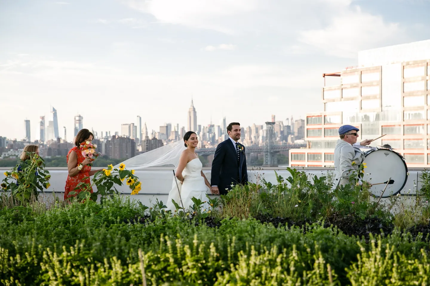 Alexis and David walk hand in hand past sunflowers, a drummer leading them out