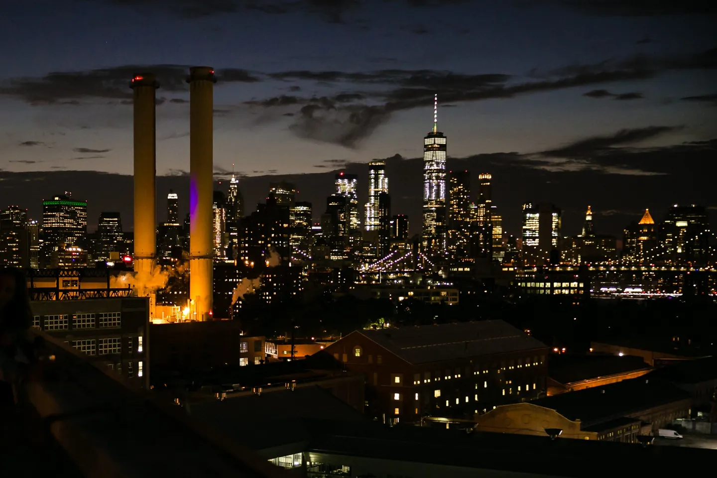 The Manhattan skyline glows at night beyond the dark rooftops of Brooklyn