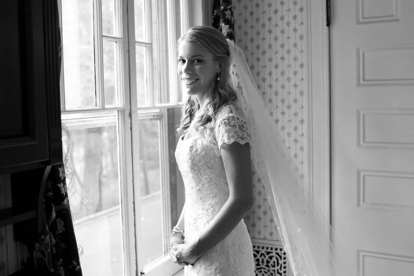 The bride stands in soft window light in a lace gown and veil, in black and white