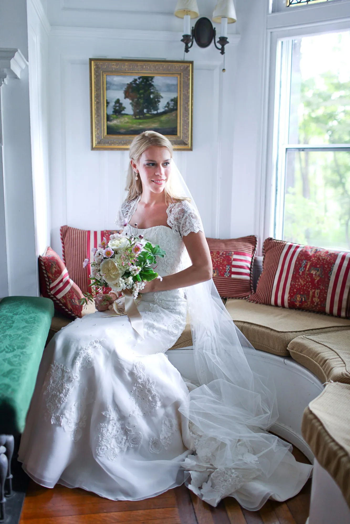 The bride sits on a cushioned window seat in a white room, bouquet in her lap