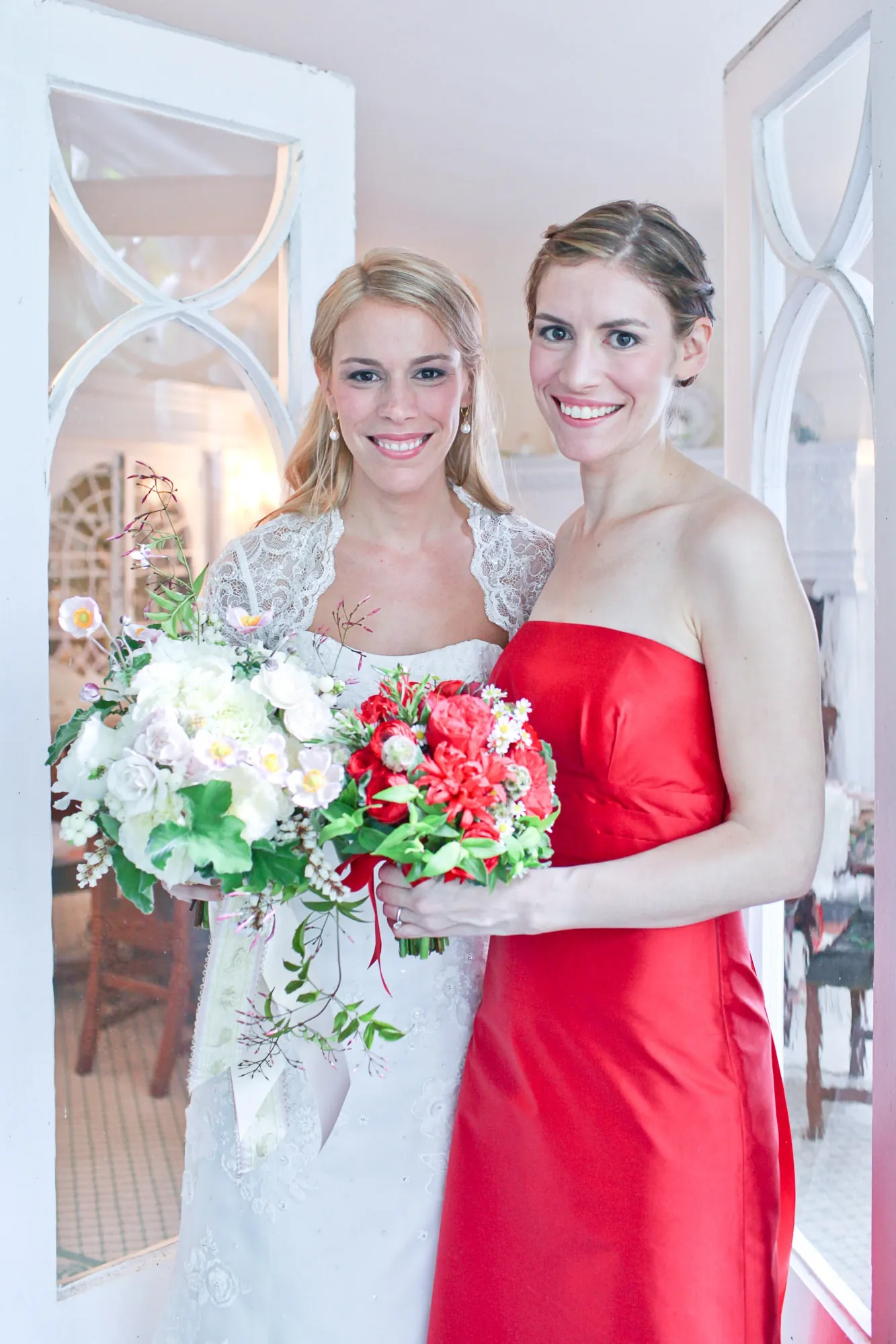 The bride and a bridesmaid in scarlet stand together in a white doorway
