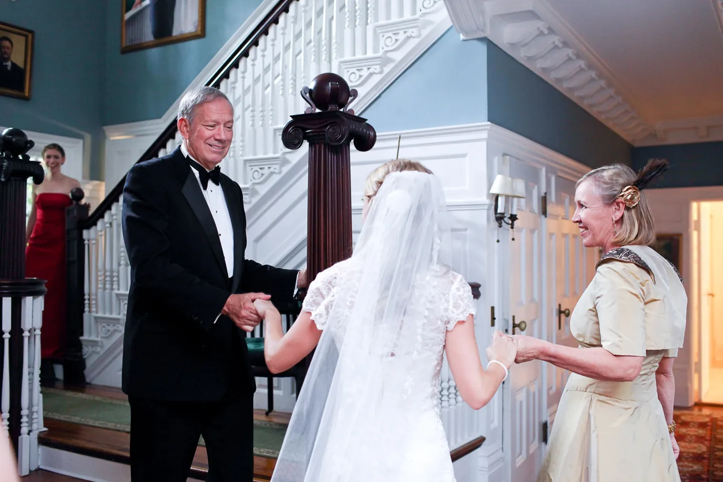 A man in a tuxedo and a woman in gold silk take the bride's hands by the staircase