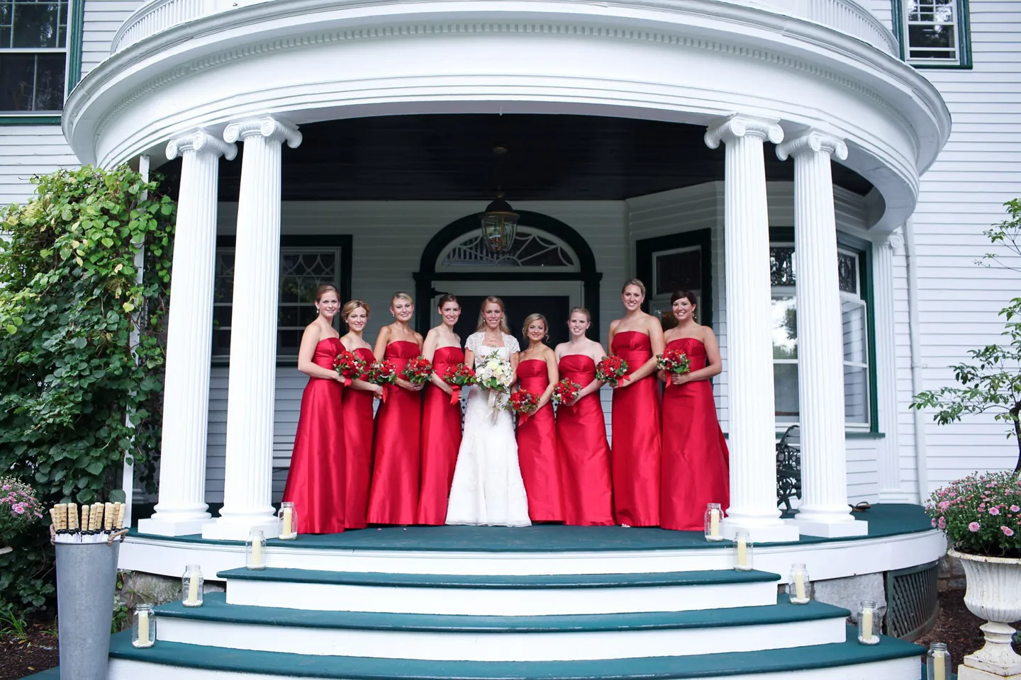 Bridesmaids in scarlet gowns stand on the columned porch of a white Victorian house