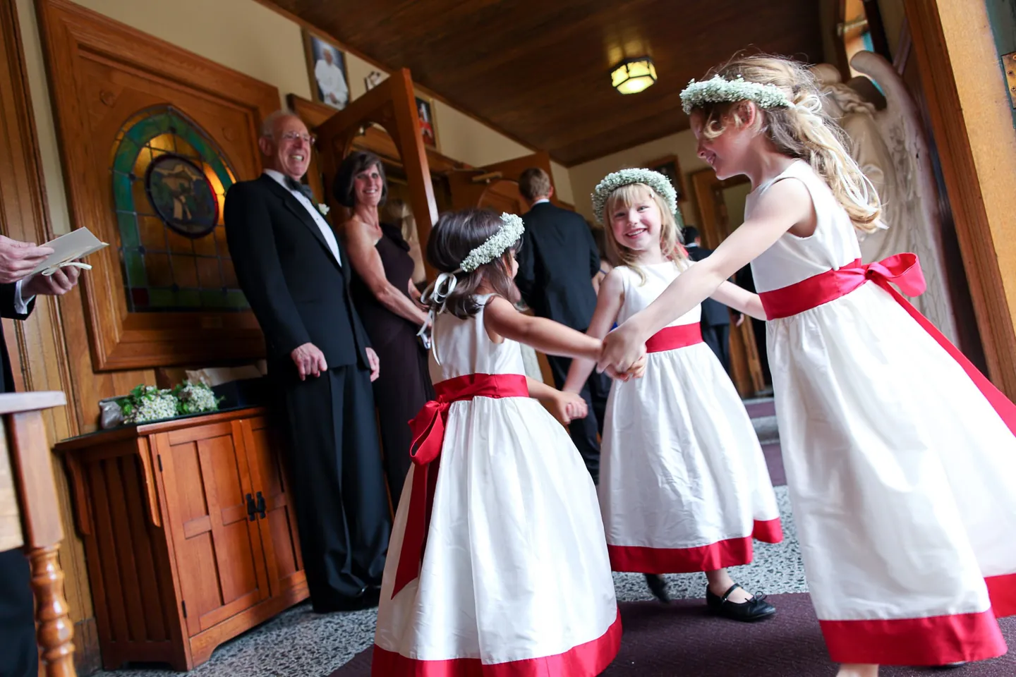 Flower girls in white dresses with red sashes wait in a wood-paneled hall