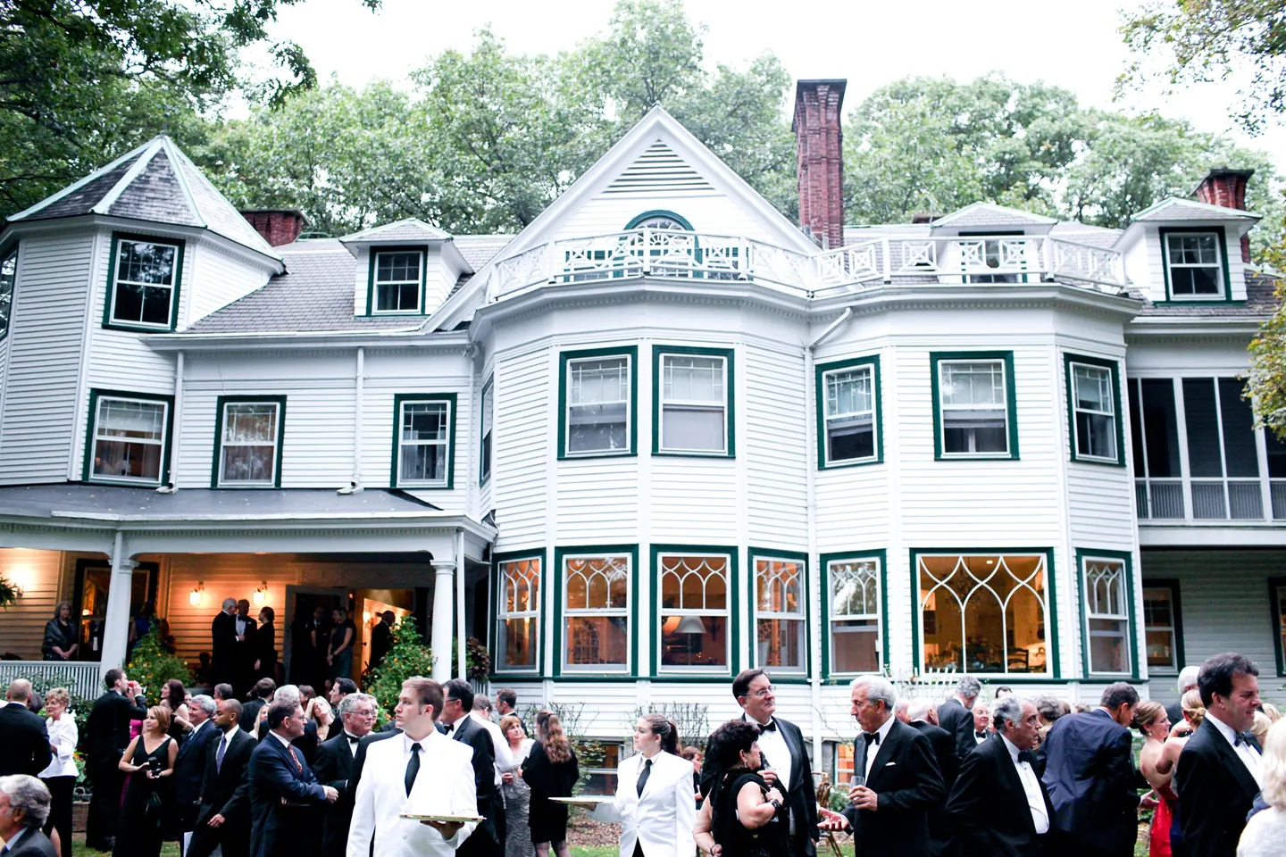 Guests mingle on the lawn in front of a large white Victorian house with a turret