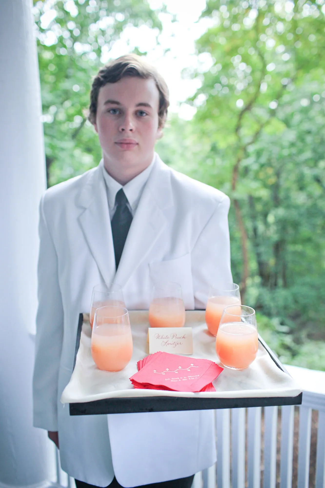 A young server in a white jacket carries a tray of orange drinks on the porch