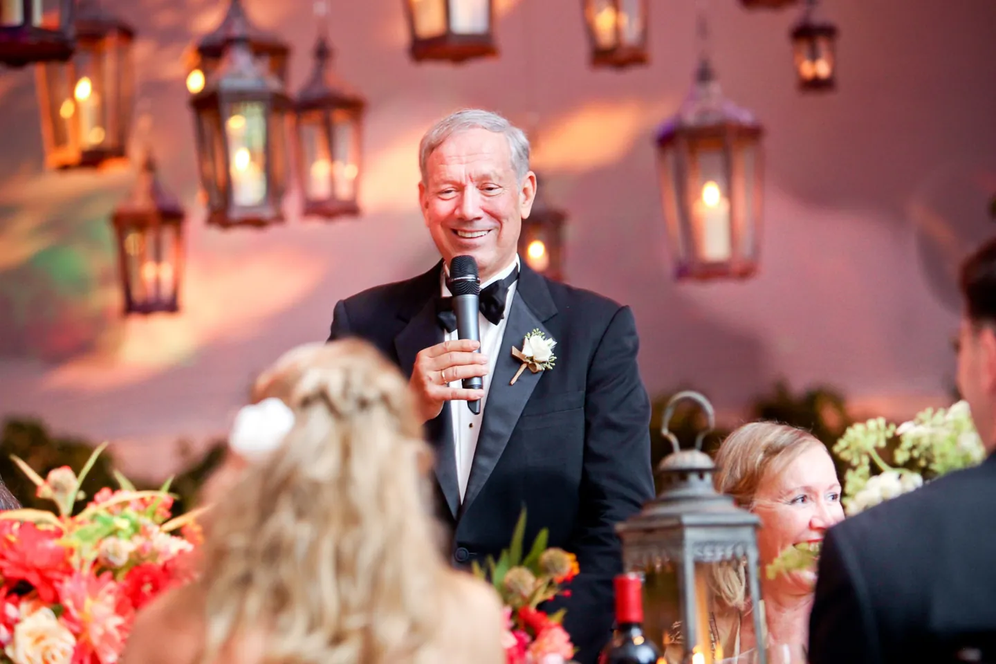 A man in a tuxedo gives a toast beneath hanging iron lanterns