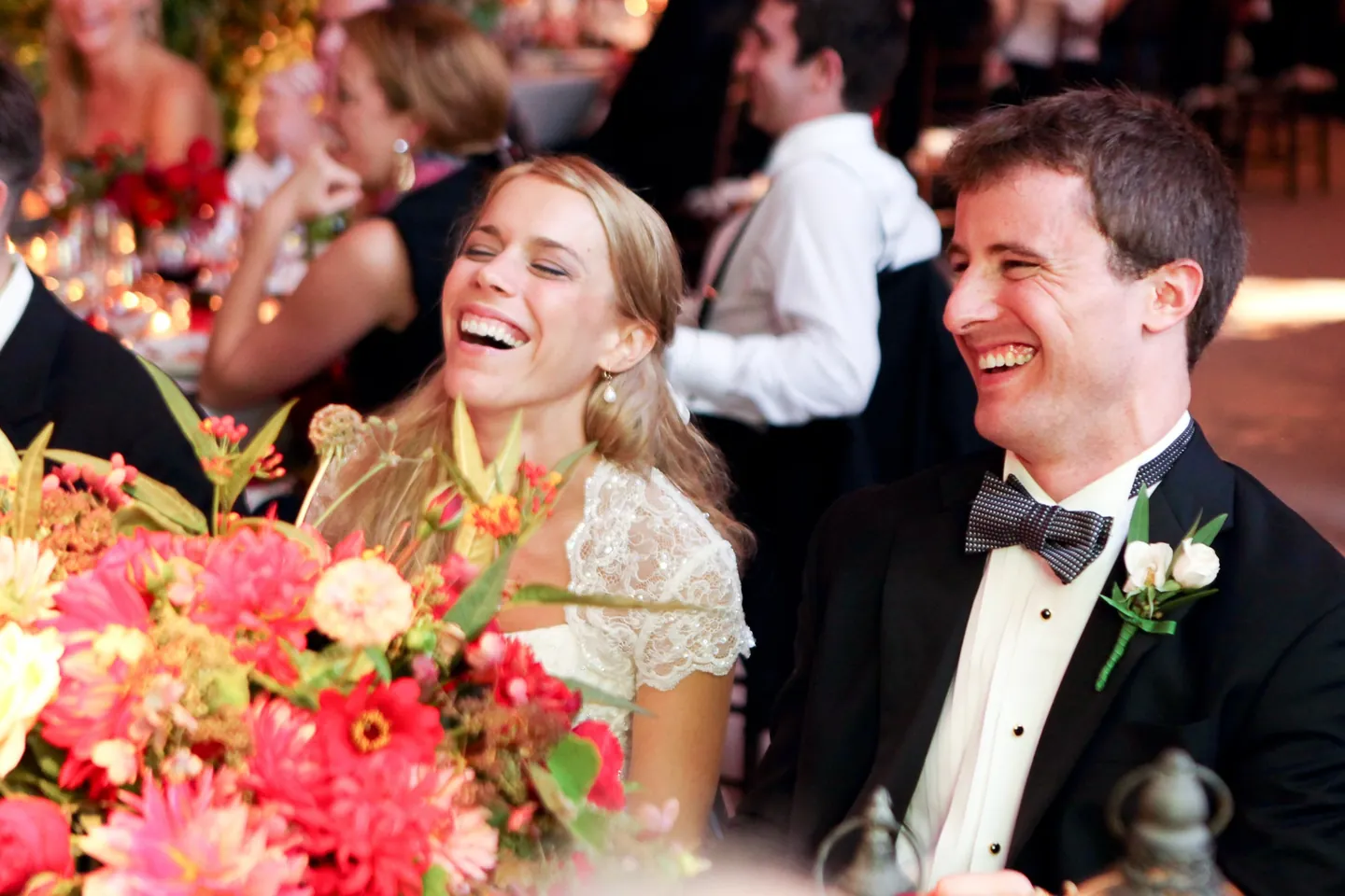 Alli and Dave laugh together at the table behind a mound of red flowers