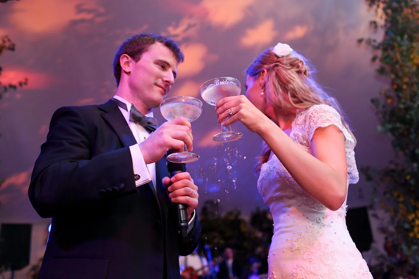 Alli and Dave link arms to sip from etched coupe glasses under purple light