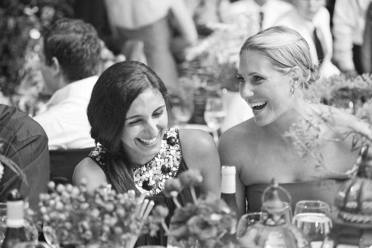 Guests laugh at a crowded dinner table, in black and white