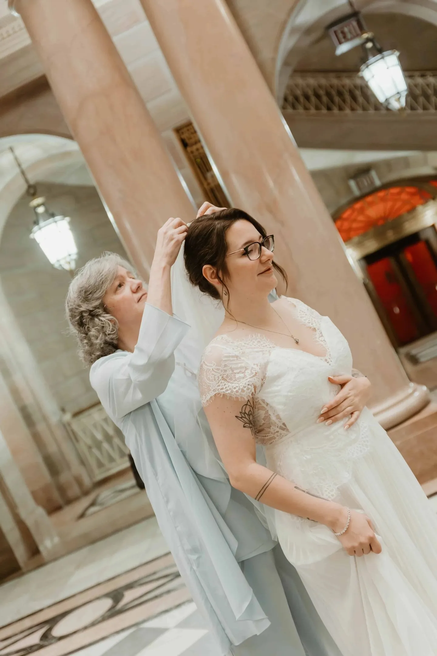 A woman in pale blue silk adjusts the bride's veil between pink marble columns