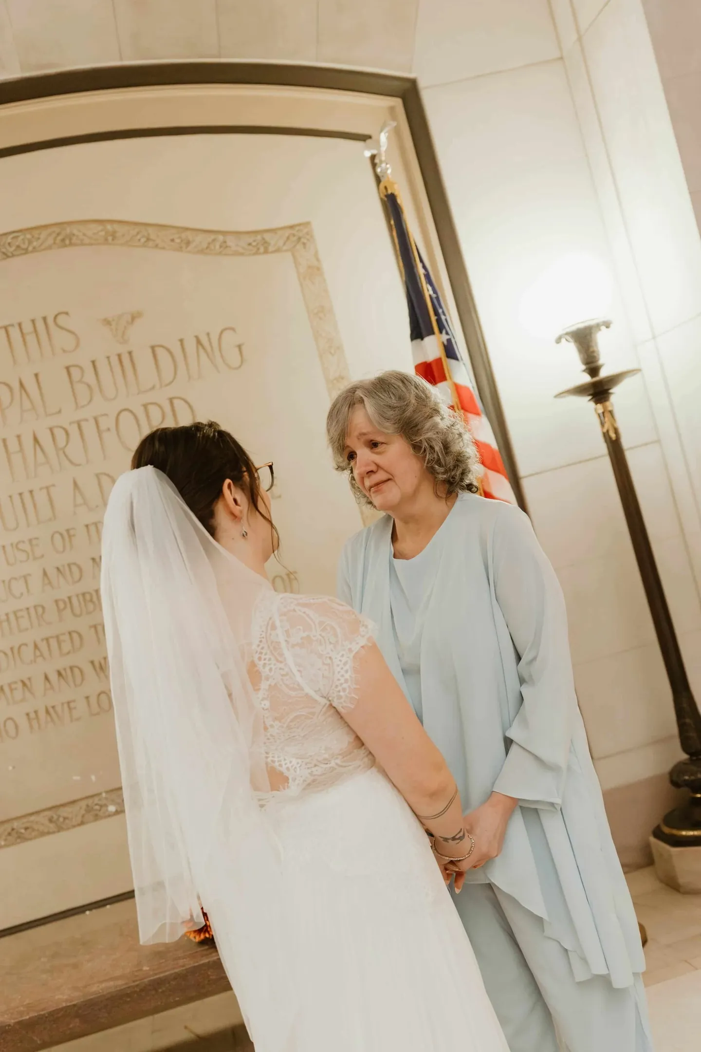 The bride faces a woman in pale blue beside an engraved stone wall and a flag