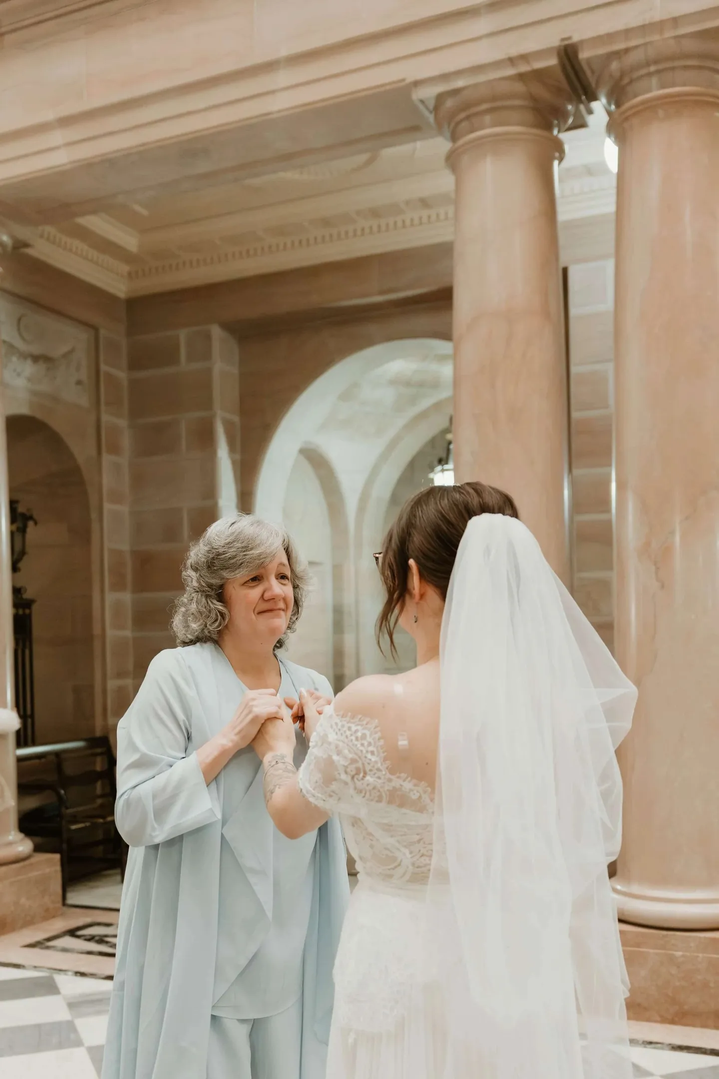 A woman in blue clasps her hands as she looks at the bride among marble columns