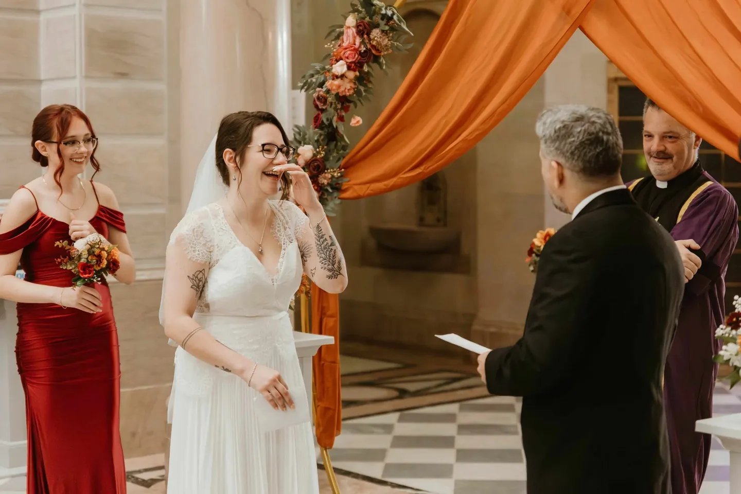 The bride laughs through tears under orange silk as the groom reads his vows from a card