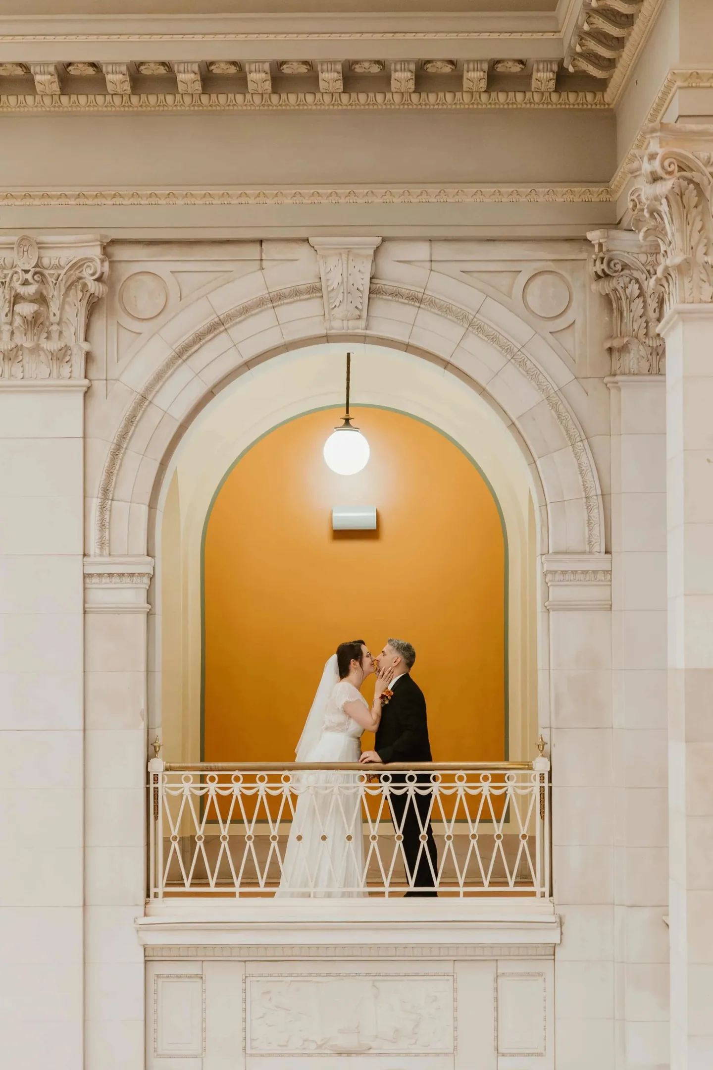 Amber and Marco kiss on a balcony framed by a glowing mustard-colored arch